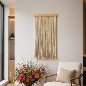 May include: A beige macrame wall hanging with a wooden top is displayed on a white wall. A cream-colored armchair with wooden arms and a beige cushion sits next to a large glass vase filled with colorful flowers.