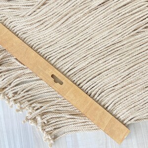 May include: A light brown wooden slat with a keyhole-shaped cutout, resting on a cream-colored macrame wall hanging with a fringed edge. The macrame is made of twisted rope.