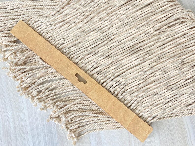 May include: A light brown wooden slat with a keyhole-shaped cutout, resting on a cream-colored macrame wall hanging with a fringed edge. The macrame is made of twisted rope.