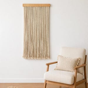 May include: A beige macrame wall hanging with a wooden top, hanging against a white wall. A cream-colored armchair with a matching pillow and a jute rug are in the foreground, creating a cozy, neutral-toned interior design.