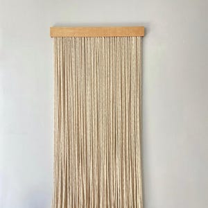 May include: A natural-toned macrame wall hanging. The piece features numerous vertical strands of off-white rope suspended from a light-colored wooden bar. The bottom of the rope strands are knotted, creating a fringe effect. This decorative item is designed for interior decor.