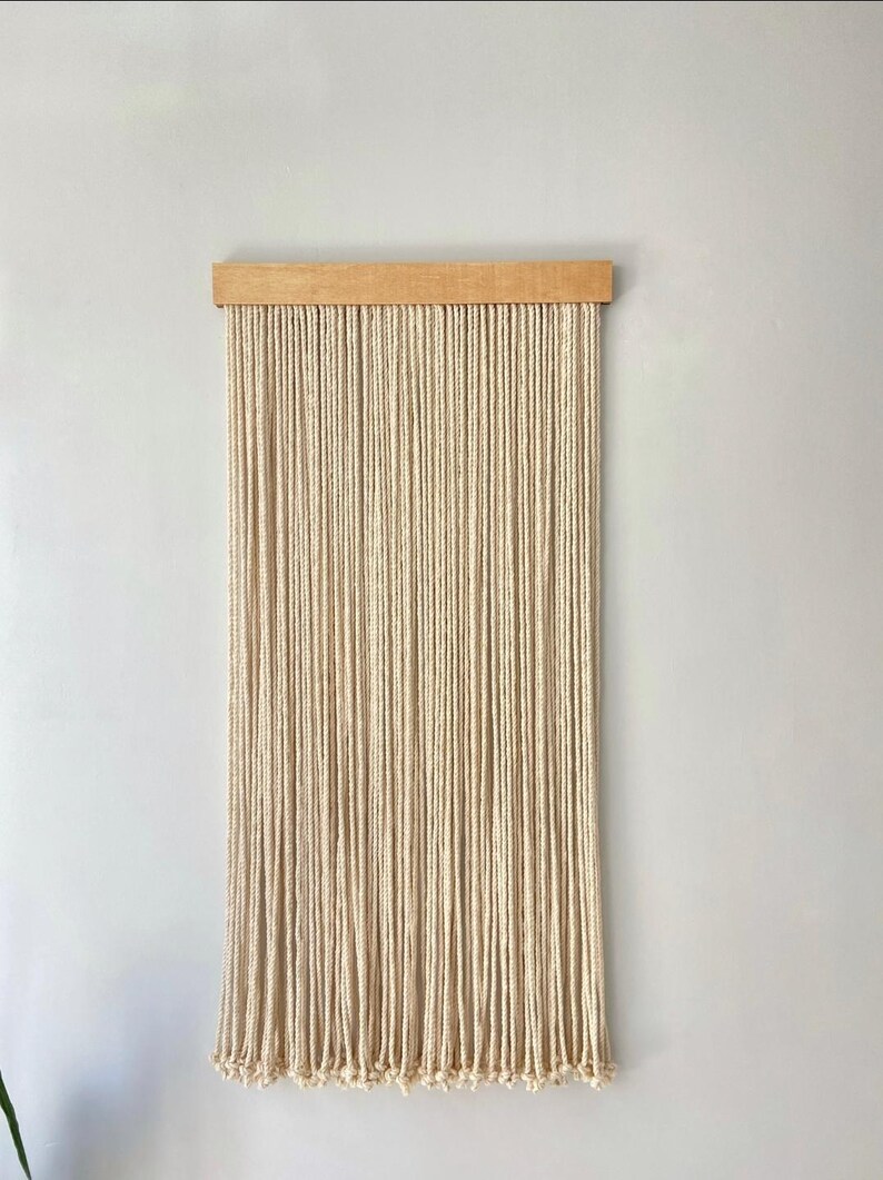 May include: A natural-toned macrame wall hanging. The piece features numerous vertical strands of off-white rope suspended from a light-colored wooden bar. The bottom of the rope strands are knotted, creating a fringe effect. This decorative item is designed for interior decor.