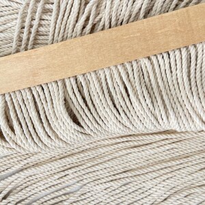 May include: Close-up of macrame cord in a natural off-white color. A light-colored wooden dowel is placed across the top of the cord. The cord is tightly twisted and appears to be ready for crafting projects.