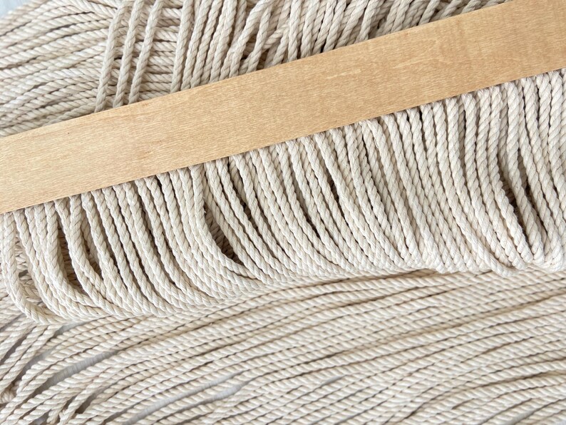 May include: Close-up of macrame cord in a natural off-white color. A light-colored wooden dowel is placed across the top of the cord. The cord is tightly twisted and appears to be ready for crafting projects.
