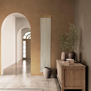 Op de afbeelding: Interieur met een neutrale kleurenpalet. Een houten kast herbergt planten en boeken. Een crèmekleurig gordijn hangt aan een gouden stang. Een grote grijze vaas staat op de vloer. Gewelfde deuropeningen leiden naar een lichte gang.
