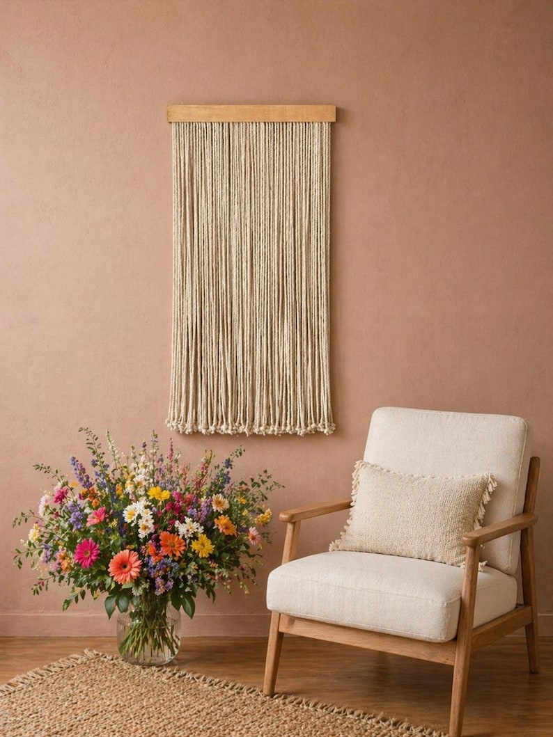 May include: A beige macrame wall hanging with a wooden top is displayed on a peach-colored wall. A cream-colored armchair with a fringed pillow sits nearby. A large vase of colorful flowers sits on a jute rug.