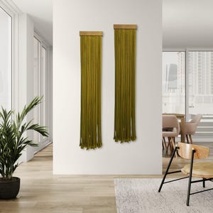 Puede incluir: Dos tapices de pared de macramé verde oliva con tapas de madera se exhiben en una pared blanca. Los largos hilos verticales del macramé crean un elemento decorativo texturizado. La habitación tiene un diseño moderno y minimalista.