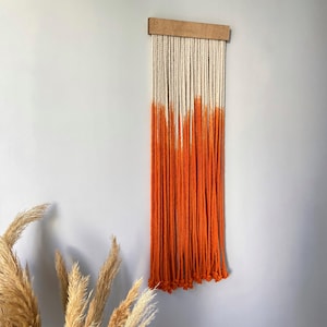 May include: A decorative wall hanging with a wooden bar and multiple rope strands. The top part of the rope is white, fading into a bright orange at the bottom. The rope is knotted at the base. This piece adds a splash of colour to any room, measuring approximately 60 cm in length.