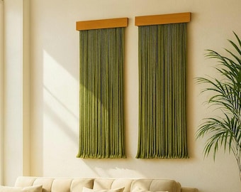 May include: Two olive green curtains with wooden valances hang on a cream-colored wall. The curtains are long and pleated, with a vertical striped texture. A large green plant is to the right of the curtains.