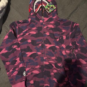Op de afbeelding: Een paarse camouflage hoodie met ritssluiting en een haaiengezicht op de capuchon. De hoodie heeft een volledige ritssluiting, geribde manchetten en een kangoeroezak. Het camouflagepatroon bevat tinten paars, roze en marineblauw.