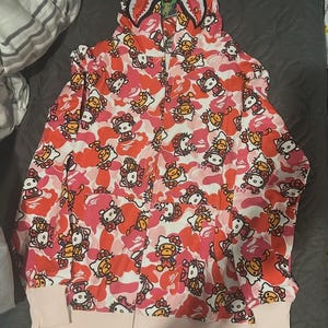 May include: Pink and red camouflage zip-up hoodie featuring a repeating pattern of Hello Kitty and ape characters. The hood has a shark-like design with a zipper that goes up to the top of the hood. The cuffs and bottom hem are a light pink color.