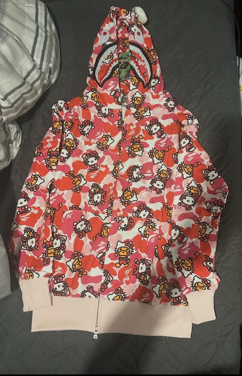 May include: Pink and red camouflage zip-up hoodie featuring a repeating pattern of Hello Kitty and ape characters. The hood has a shark-like design with a zipper that goes up to the top of the hood. The cuffs and bottom hem are a light pink color.