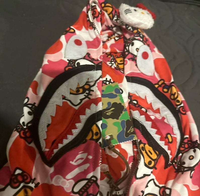May include: A pink, red, and white zip-up hoodie with a shark mouth design and Hello Kitty accents. The hoodie features a camouflage tag and a white Hello Kitty plush attached to the hood.