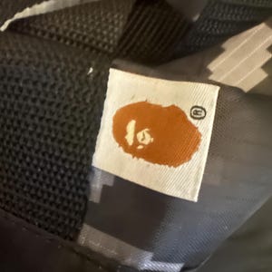 May include: Close-up of a white tag with a brown ape head logo and a small, circular registered trademark symbol. The tag is sewn onto a black and gray camouflage-patterned fabric, likely part of a bag or accessory.