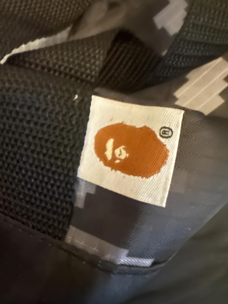 May include: Close-up of a white tag with a brown ape head logo and a small, circular registered trademark symbol. The tag is sewn onto a black and gray camouflage-patterned fabric, likely part of a bag or accessory.