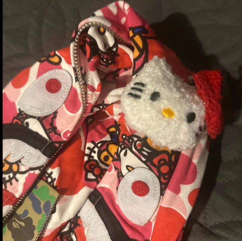 May include: A pink and red zip-up hoodie with a repeating pattern of cartoon characters and white accents. A white plush Hello Kitty head with a red bow is attached to the hoodie. The hoodie has a silver zipper.