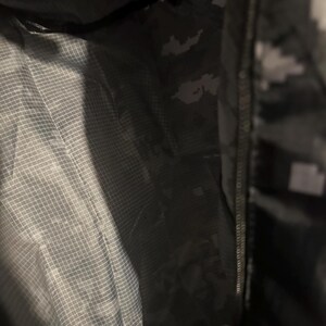 May include: Interior view of a dark green and gray camouflage jacket. The jacket has a zipper closure and a light gray, grid-patterned lining. The camouflage pattern is visible on the outer fabric.