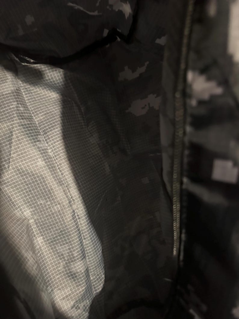 May include: Interior view of a dark green and gray camouflage jacket. The jacket has a zipper closure and a light gray, grid-patterned lining. The camouflage pattern is visible on the outer fabric.