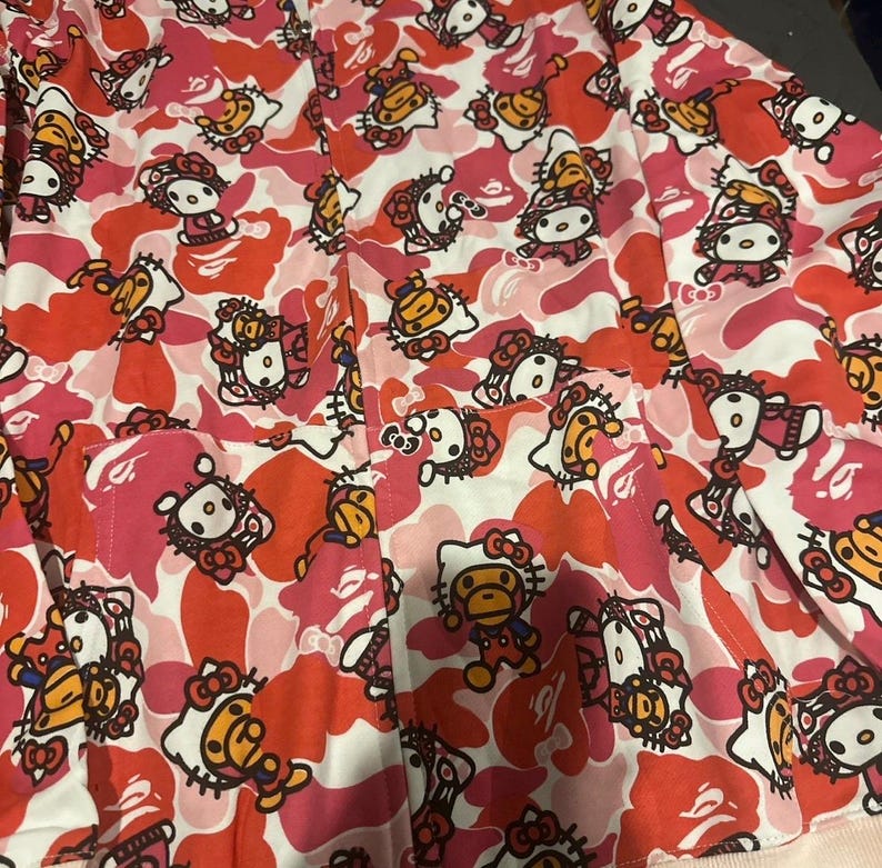 May include: A pink, red, and white patterned button-up shirt. The pattern features a camouflage design with cartoon characters. The shirt has a pocket on the front and a collar.