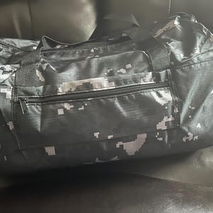 May include: A black and white digital camouflage duffel bag with a zippered front pocket and black handles. The bag is made of a durable material and is designed for travel or sports.