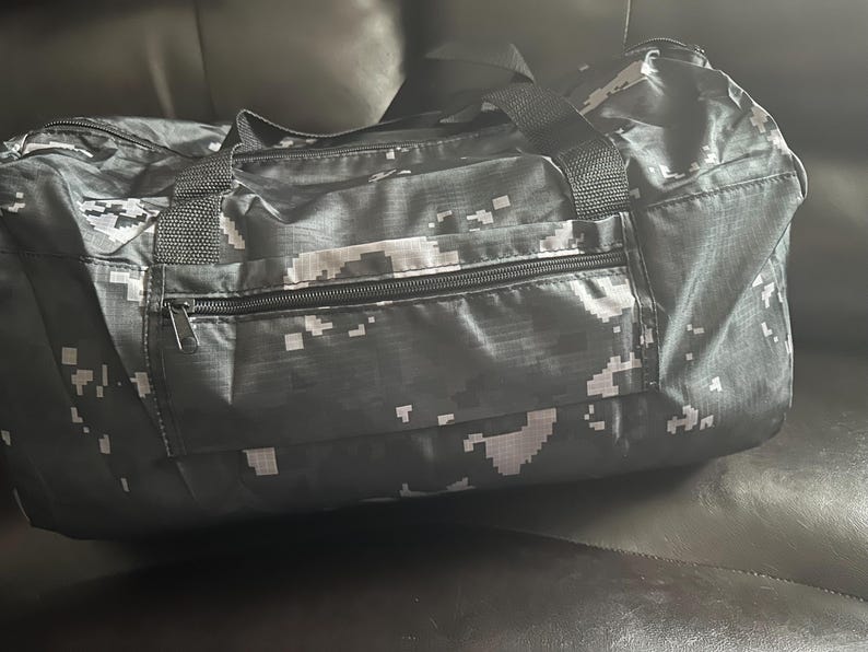 May include: A black and white digital camouflage duffel bag with a zippered front pocket and black handles. The bag is made of a durable material and is designed for travel or sports.