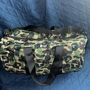 May include: A camouflage-patterned duffel bag with black handles and a shoulder strap. The bag features a green, brown, and tan camouflage design. The brand name "BAPE" is visible on the front of the bag.