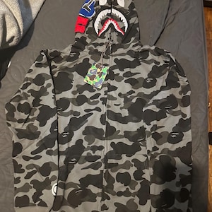 Puede incluir: Una sudadera con capucha y cremallera completa con un patrón de camuflaje en gris oscuro y negro. La capucha muestra un gráfico de boca de tiburón con dientes rojos, blancos y negros, y un parche azul, naranja y rojo en el lateral.