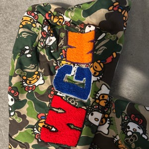Puede incluir: Una sudadera con capucha con estampado de camuflaje y un dise&ntilde;o repetido de personajes de dibujos animados. La sudadera tiene letras grandes y coloridas en naranja, azul y rojo. El dise&ntilde;o combina elementos l&uacute;dicos con una est&eacute;tica de inspiraci&oacute;n militar.