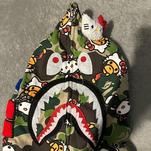 Puede incluir: Una sudadera con capucha con cremallera y estampado de camuflaje con un dise&ntilde;o de cara de tibur&oacute;n en la capucha. La sudadera tiene una boca de tibur&oacute;n blanca y roja, ojos grandes y un peque&ntilde;o peluche de Hello Kitty adjunto a la capucha.