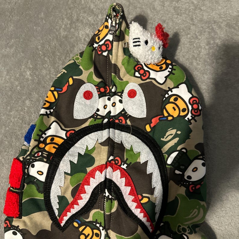 Puede incluir: Una sudadera con capucha con cremallera y estampado de camuflaje con un dise&ntilde;o de cara de tibur&oacute;n en la capucha. La sudadera tiene una boca de tibur&oacute;n blanca y roja, ojos grandes y un peque&ntilde;o peluche de Hello Kitty adjunto a la capucha.