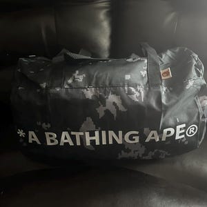 May include: A black and gray camouflage duffel bag with the text "A BATHING APE" in white. The bag has two black handles and a small tag. The bag is made of a durable material.