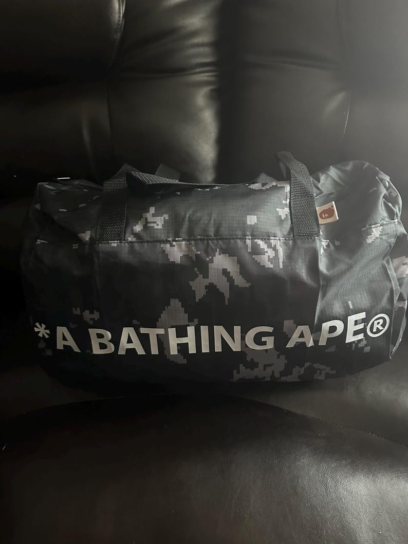 May include: A black and gray camouflage duffel bag with the text "A BATHING APE" in white. The bag has two black handles and a small tag. The bag is made of a durable material.