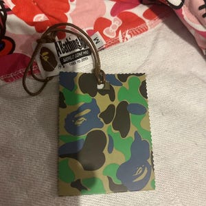 May include: A tag with a camouflage pattern in green, blue, and brown, attached to a garment with a pink and white pattern. The tag reads "A Bathing Ape" and "WORLD GONE MFG 1993 TO 2093".