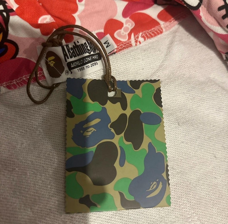 May include: A tag with a camouflage pattern in green, blue, and brown, attached to a garment with a pink and white pattern. The tag reads "A Bathing Ape" and "WORLD GONE MFG 1993 TO 2093".