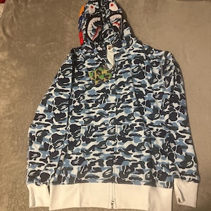 Pu&ograve; includere: Una felpa con cappuccio con zip mimetica blu e bianca con un design a tema squalo sul cappuccio. La felpa con cappuccio presenta una base bianca con motivi mimetici blu e neri. Il cappuccio ha un design a forma di faccia di squalo con accenti rossi, bianchi e blu.
