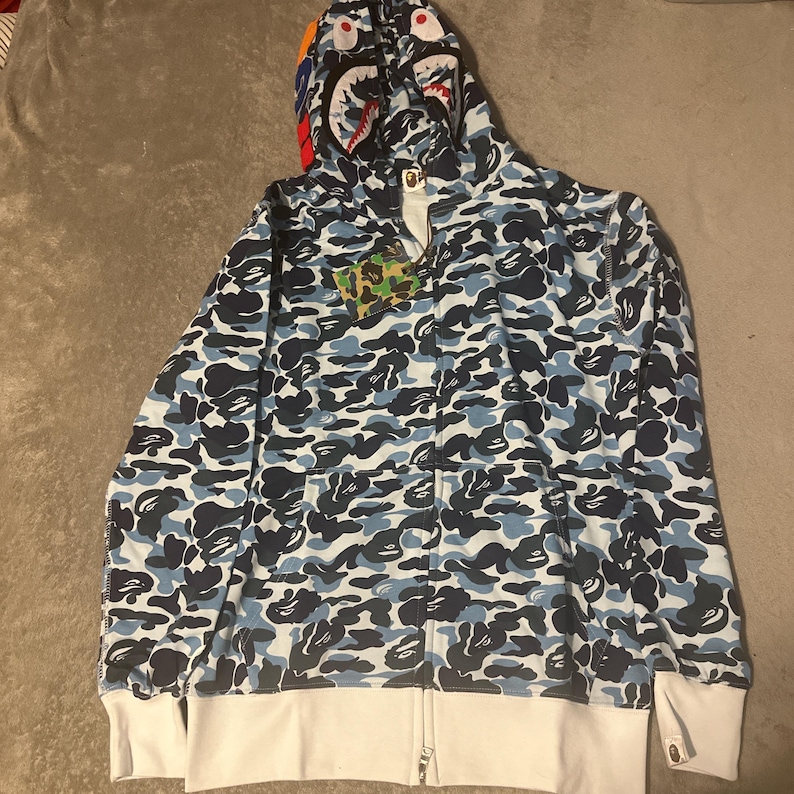 Pu&ograve; includere: Una felpa con cappuccio con zip mimetica blu e bianca con un design a tema squalo sul cappuccio. La felpa con cappuccio presenta una base bianca con motivi mimetici blu e neri. Il cappuccio ha un design a forma di faccia di squalo con accenti rossi, bianchi e blu.