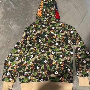 Puede incluir: Una sudadera con capucha de camuflaje verde con un patr&oacute;n de personajes de Hello Kitty. La capucha tiene detalles de rayas de tigre y rojos. Las mangas tienen pu&ntilde;os beige. La sudadera con capucha est&aacute; hecha de un material suave y c&oacute;modo.