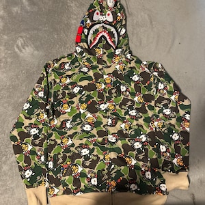 Puede incluir: Sudadera con capucha y cremallera de camuflaje verde con un patr&oacute;n repetido de Hello Kitty. La capucha presenta un dise&ntilde;o de cara de tibur&oacute;n con detalles rojos, blancos y azules. Los pu&ntilde;os y el dobladillo son de color beige.