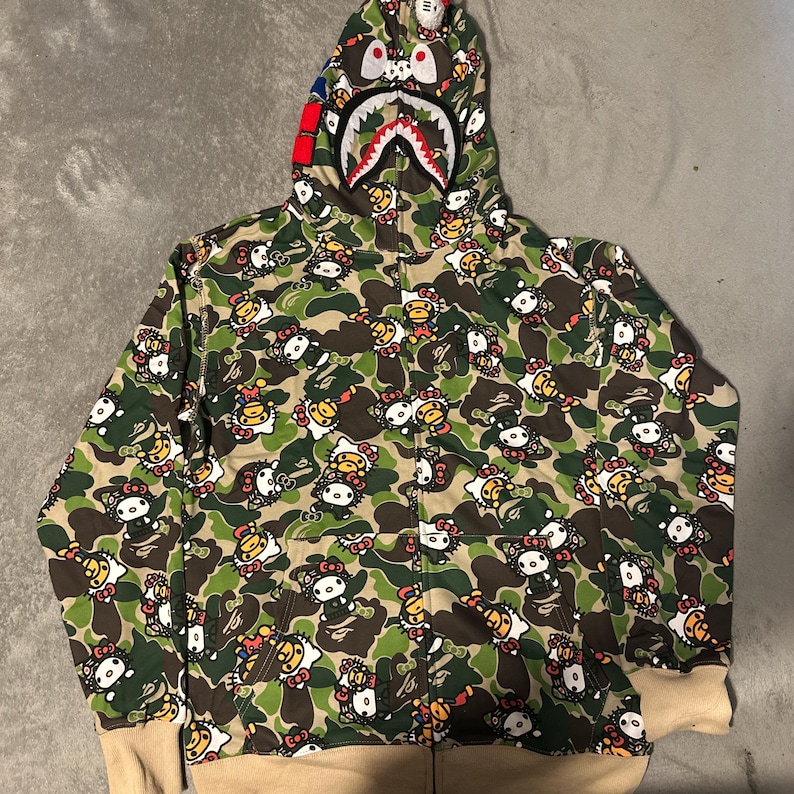 Puede incluir: Sudadera con capucha y cremallera de camuflaje verde con un patr&oacute;n repetido de Hello Kitty. La capucha presenta un dise&ntilde;o de cara de tibur&oacute;n con detalles rojos, blancos y azules. Los pu&ntilde;os y el dobladillo son de color beige.