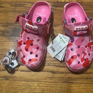 May include: Pair of pink Crocs clogs with a camouflage pattern in shades of pink and red. The clogs have a strap and the brand name "Crocs" on the strap. Includes a small bag of decorative charms.