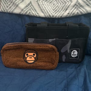 May include: A brown fuzzy pencil case with a monkey face logo and a black tote bag with a camouflage pattern and a BAPE logo. The items are on a blue surface.
