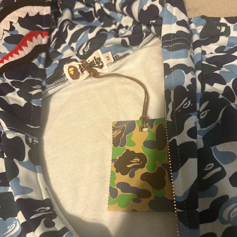 Pu&ograve; includere: Una felpa con cappuccio con zip mimetica blu, bianca e nera con un design a bocca di squalo sul cappuccio. La felpa ha un'etichetta con un motivo mimetico e il nome del marchio. L'interno &egrave; bianco.