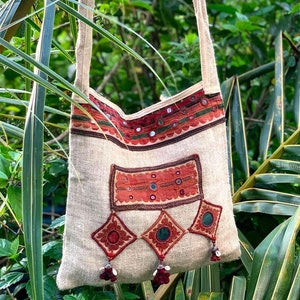 May include: A beige canvas tote bag with colourful embroidered details and small mirrors. The bag has a long strap and is decorated with red, orange, and green accents.