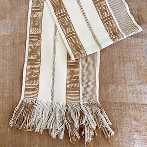 May include: Cream and brown woven scarf with a striped pattern and decorative fringe. The scarf features alternating vertical stripes of cream and brown, with detailed geometric designs in the brown stripes. The fringe at the ends adds a textured finish.
