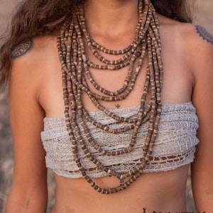 May include: A woman wearing a white crocheted top with a long layered necklace made of brown wooden beads. The necklace is made of multiple strands of beads and is worn around the neck. The woman is also wearing a belly button ring.