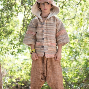 Puede incluir: Una persona joven lleva una chaqueta con capucha de rayas marrones y beige con una capucha puntiaguda. La chaqueta tiene cierre de botones y bolsillos. La persona lleva pantalones marrones y está de pie en un entorno boscoso.