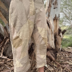 May include: Tan patchwork capri pants with a button closure and belt loops. The capri pants feature a variety of tan and beige fabric patches. The pants are knee-length and have a relaxed fit.