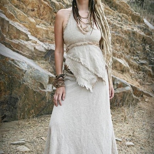 May include: A beige halter-neck dress with a layered, asymmetrical top and a flowing skirt. The dress has a raw, textured appearance, suggesting a natural fabric. The model is wearing multiple beaded necklaces and bracelets, with long dreadlocks.