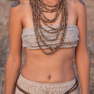 May include: A woman wearing a beige top and pants with a brown beaded necklace and a brown beaded belt.