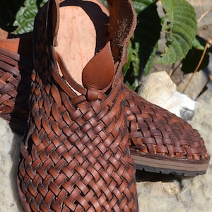 May include: A pair of brown leather sandals with a woven design. The sandals have a strap that goes around the ankle and a buckle closure.